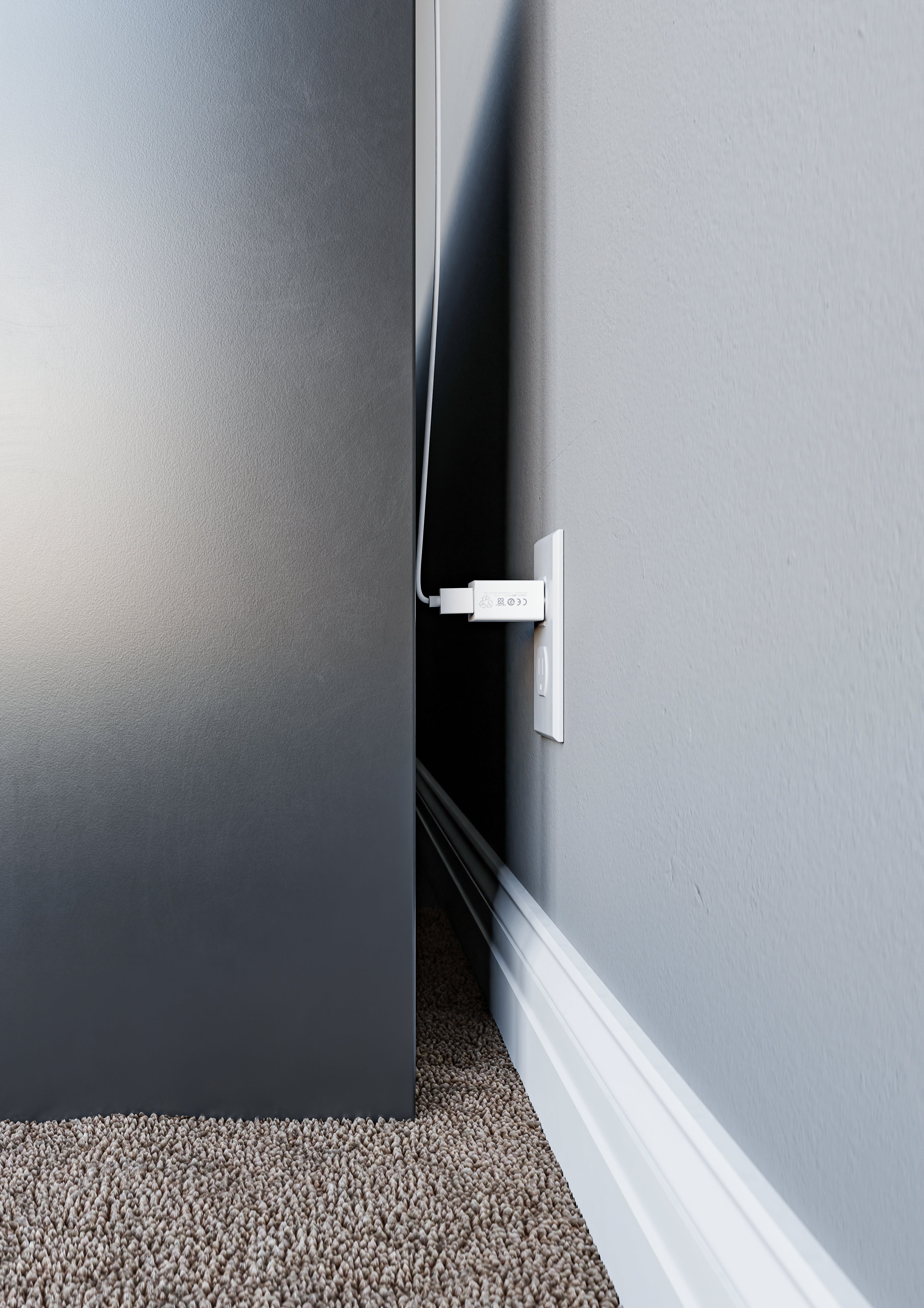 This image shows a close-up of a white charging adapter plugged into a wall outlet, positioned behind a large piece of furniture, possibly a desk or cabinet. The charging cable is routed upwards along the wall, while the outlet is barely visible due to the tight space between the furniture and the wall. The setup highlights the challenge of accessing outlets in tight spaces.