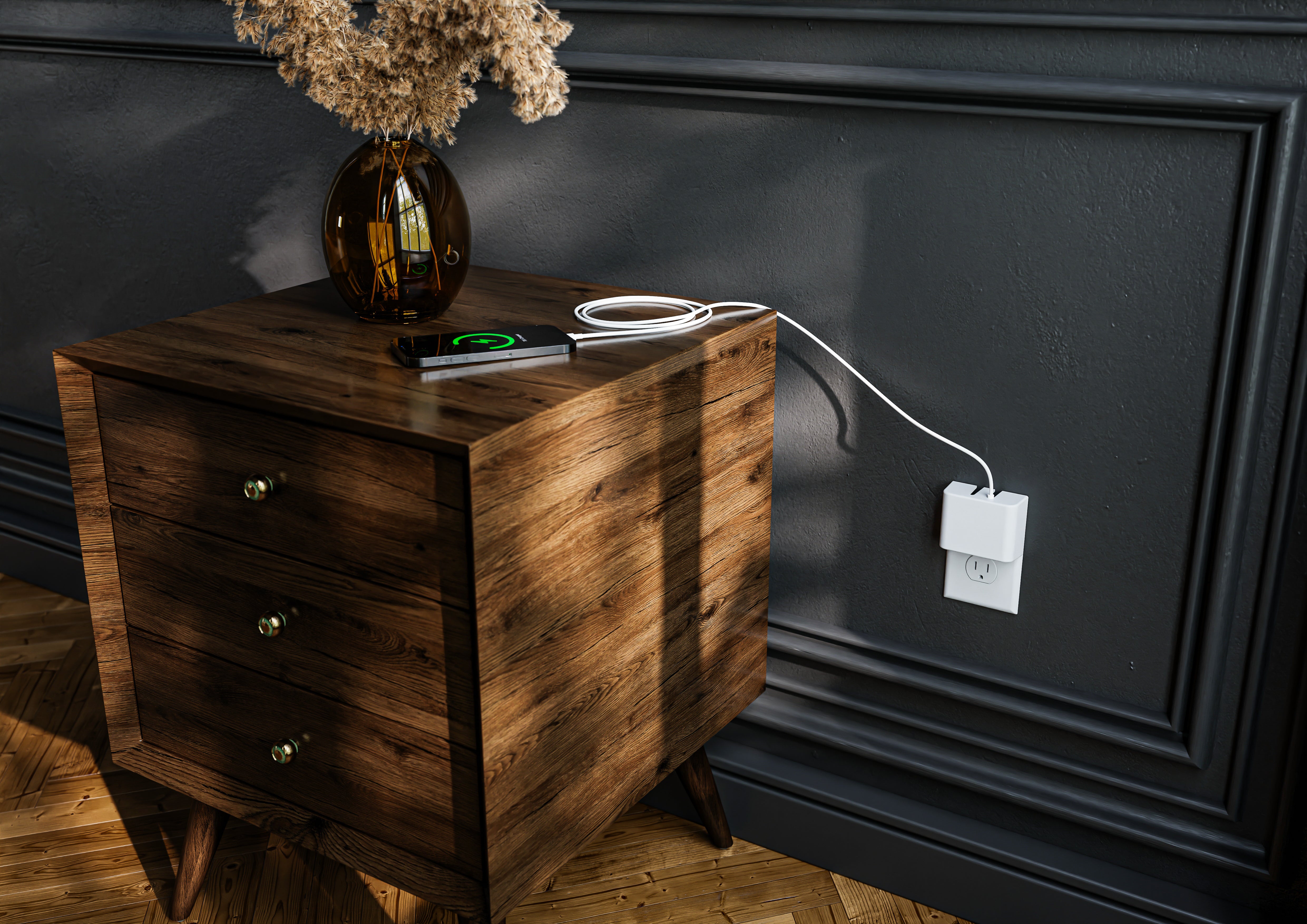 This image shows the Permaplug Charger Lock plugged into a wall outlet near a rustic wooden bedside table. The charger is powering a smartphone, which is wirelessly charging on the table, alongside a neatly coiled charging cable. A decorative vase with dried flowers adds a touch of style to the cozy and organized bedroom setting, illuminated by natural sunlight.